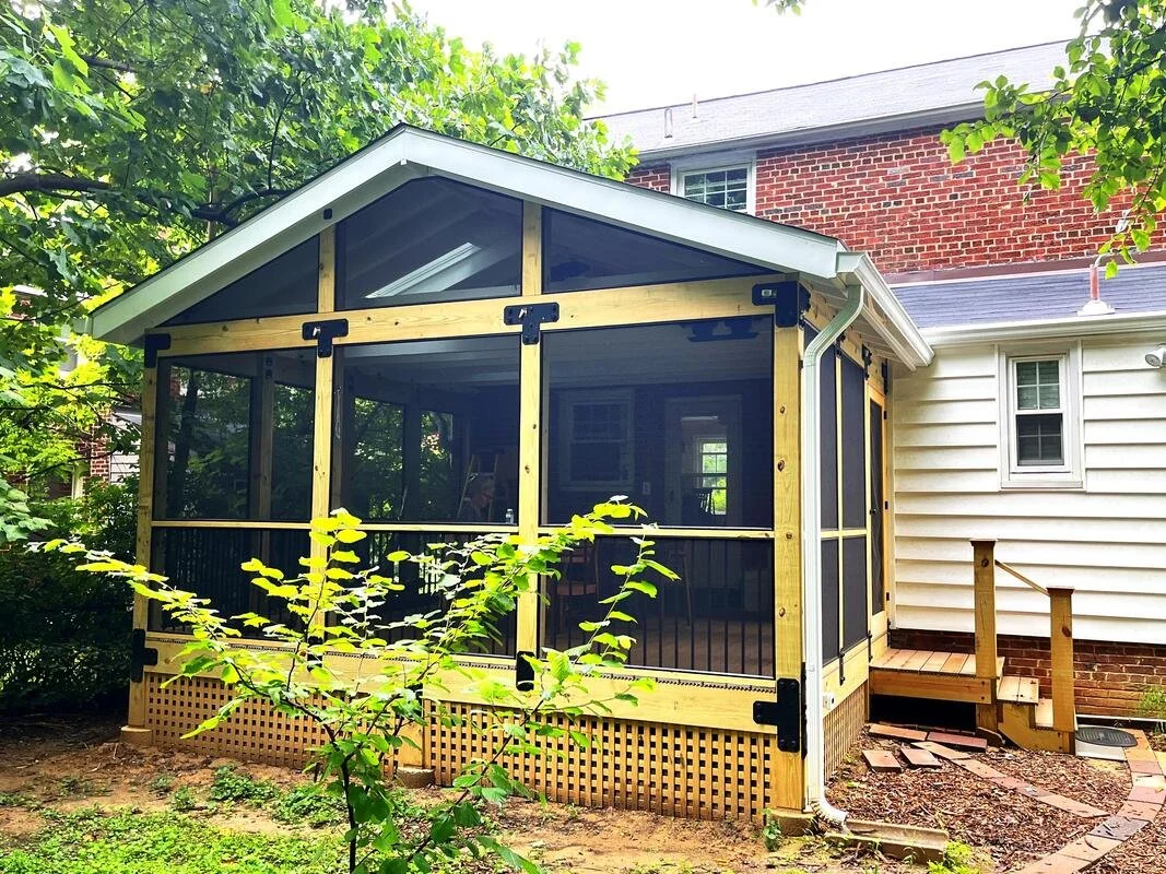 A screened-in porch or extension being built on the back of a house, with a wooden frame and black hardware, surrounded by green plants and trees.