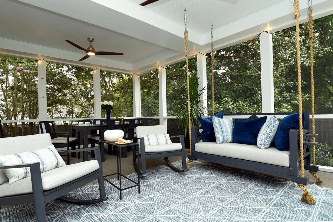 Porch with white ceiling, ceiling fan, string lights, black dining table with chairs, white sofas with blue cushions, potted plants, and a hanging swing with ropes, overlooking trees.