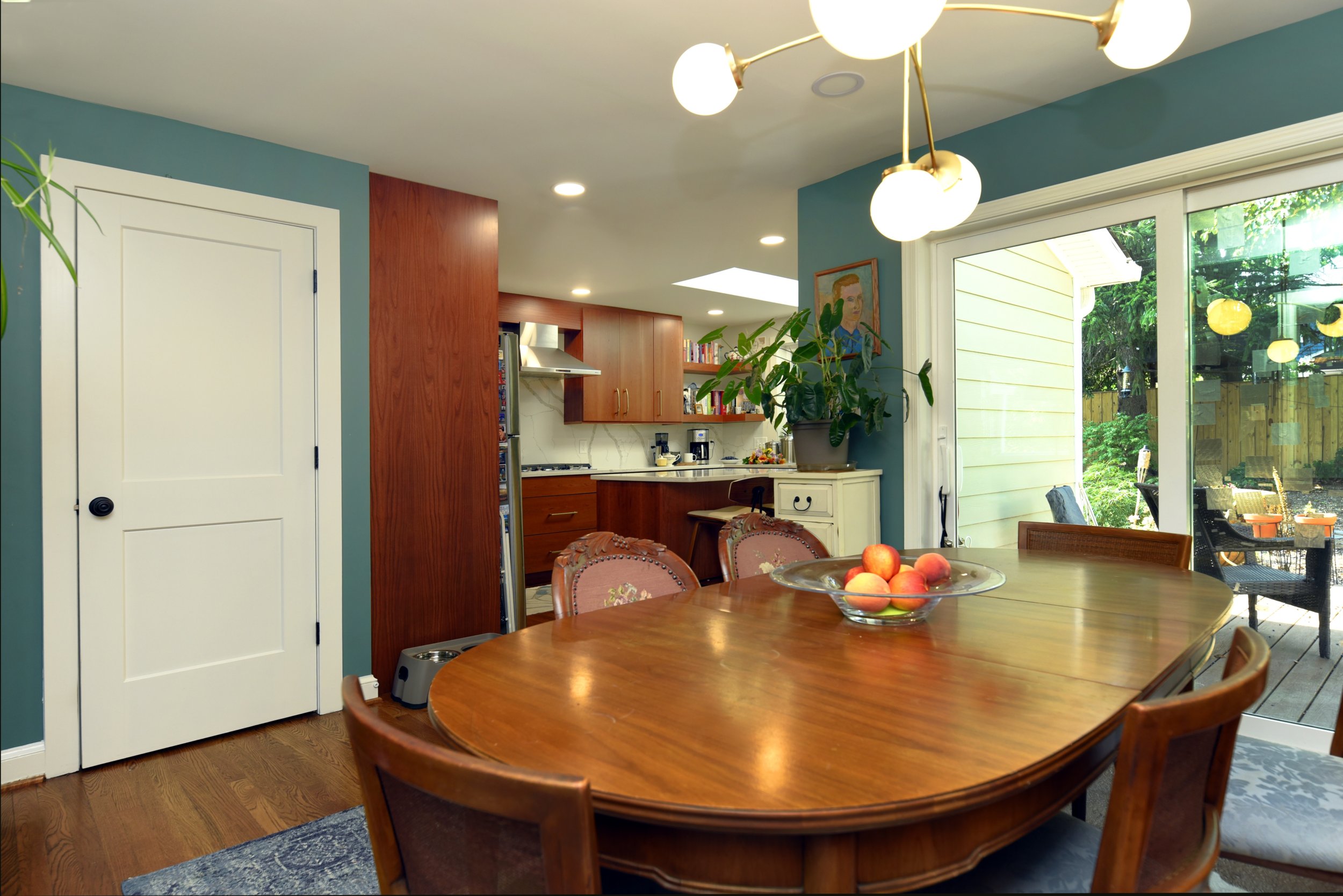 Dining room with wooden table, fruit bowl, hanging light fixture, large sliding glass door, and kitchen in the background.