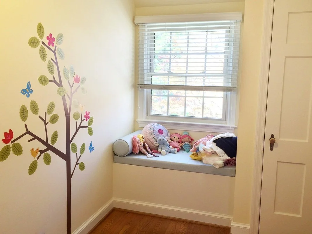 A small window nook with a cushioned seat holding stuffed animals and clothes, next to a wall decorated with a colorful leaf and animal sticker.