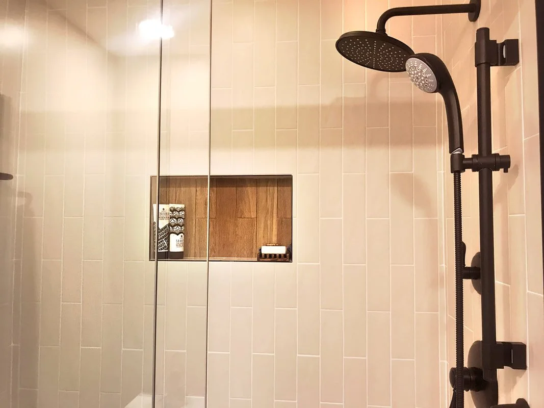 Shower with beige tiles, black showerhead and controls, built-in wooden shelf with toiletries.