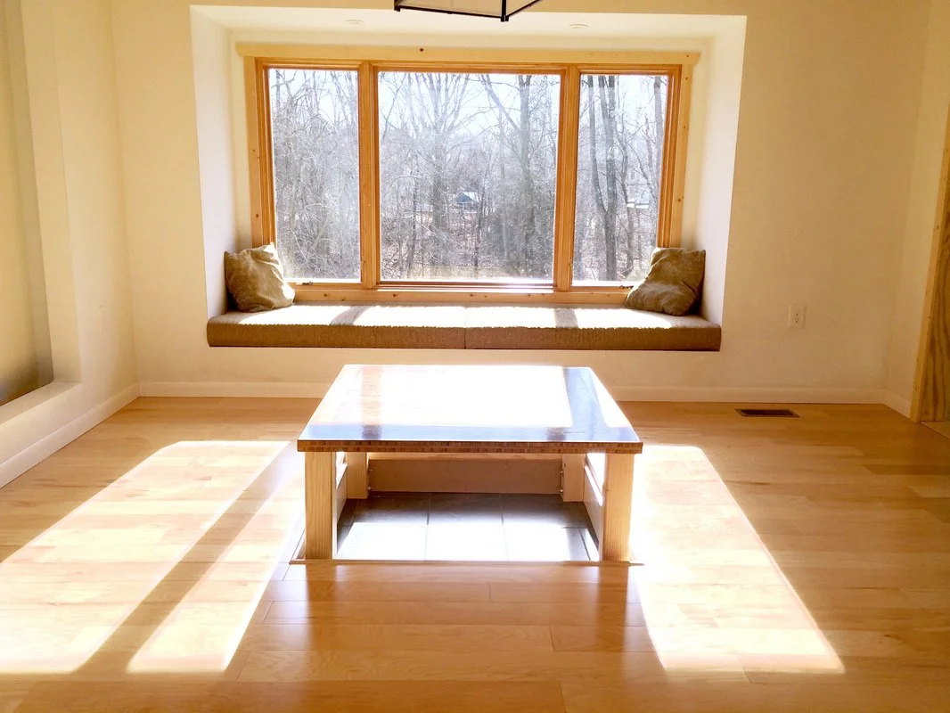 Sunlit room with wooden floors, a large window seat with beige cushions, and a coffee table in front.