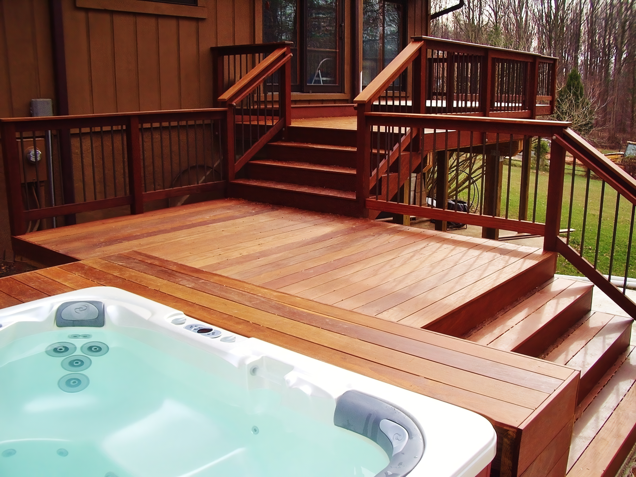 A wooden deck with stairs leading up to a house with a window, surrounded by trees. There is a hot tub filled with water and jets in the foreground.