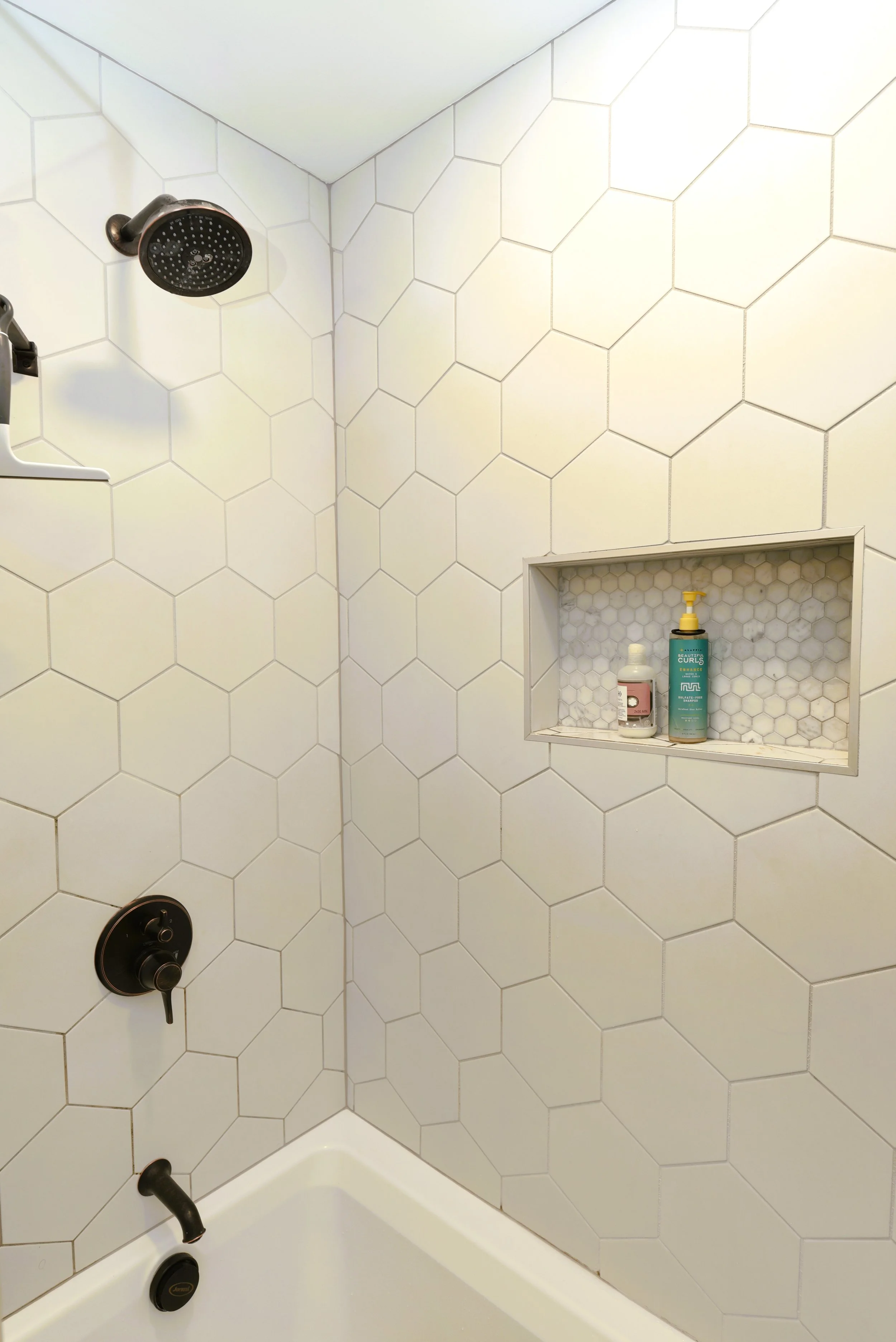 Shower area with black fixtures, white hexagonal wall tiles, and a built-in tile shelf with shampoo and conditioner bottles.