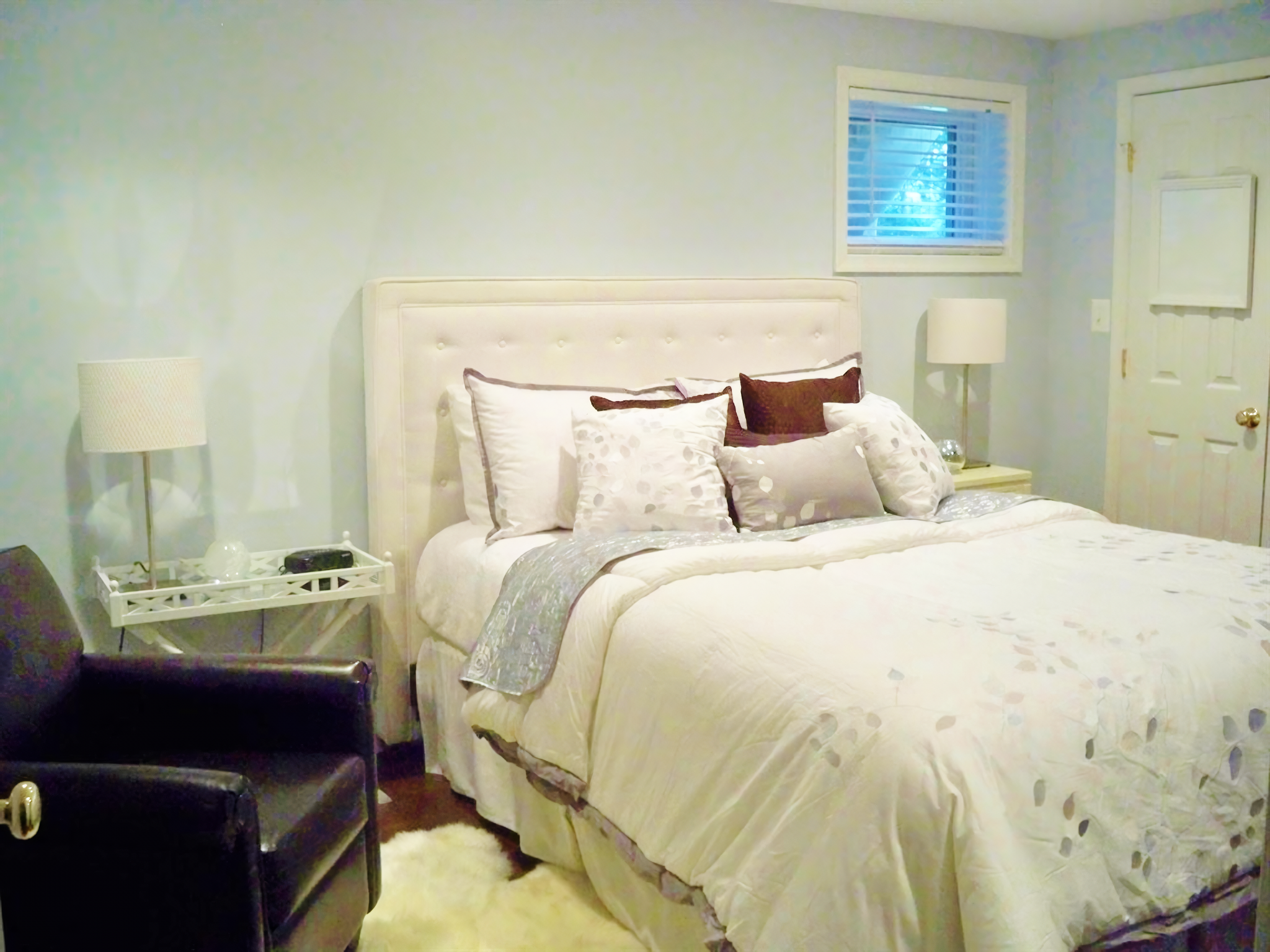 A cozy bedroom with a white upholstered bed, multiple decorative pillows, two nightstands with lamps, a window with blinds, a white door, a black armchair, and a white fluffy rug.