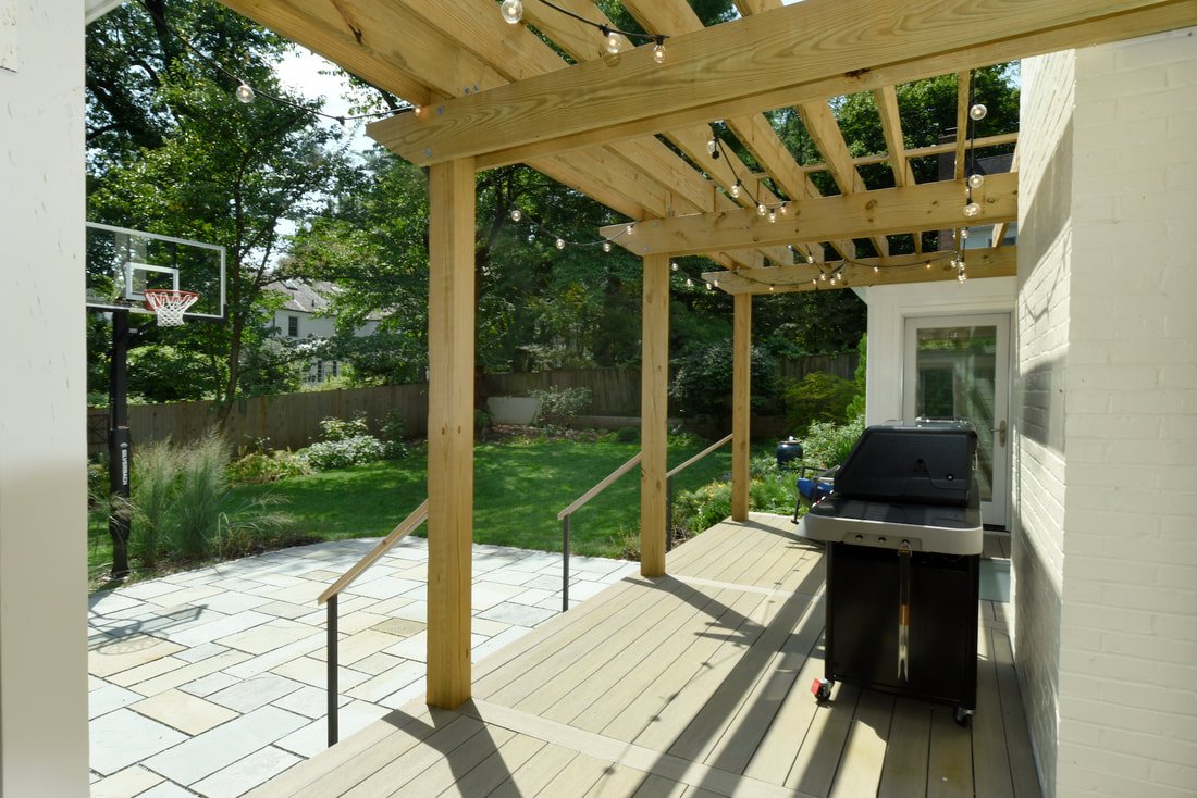 Backyard patio with a wooden pergola, string lights, a barbecue grill, a basketball hoop, a paved area, and green lawn with trees and garden plants.