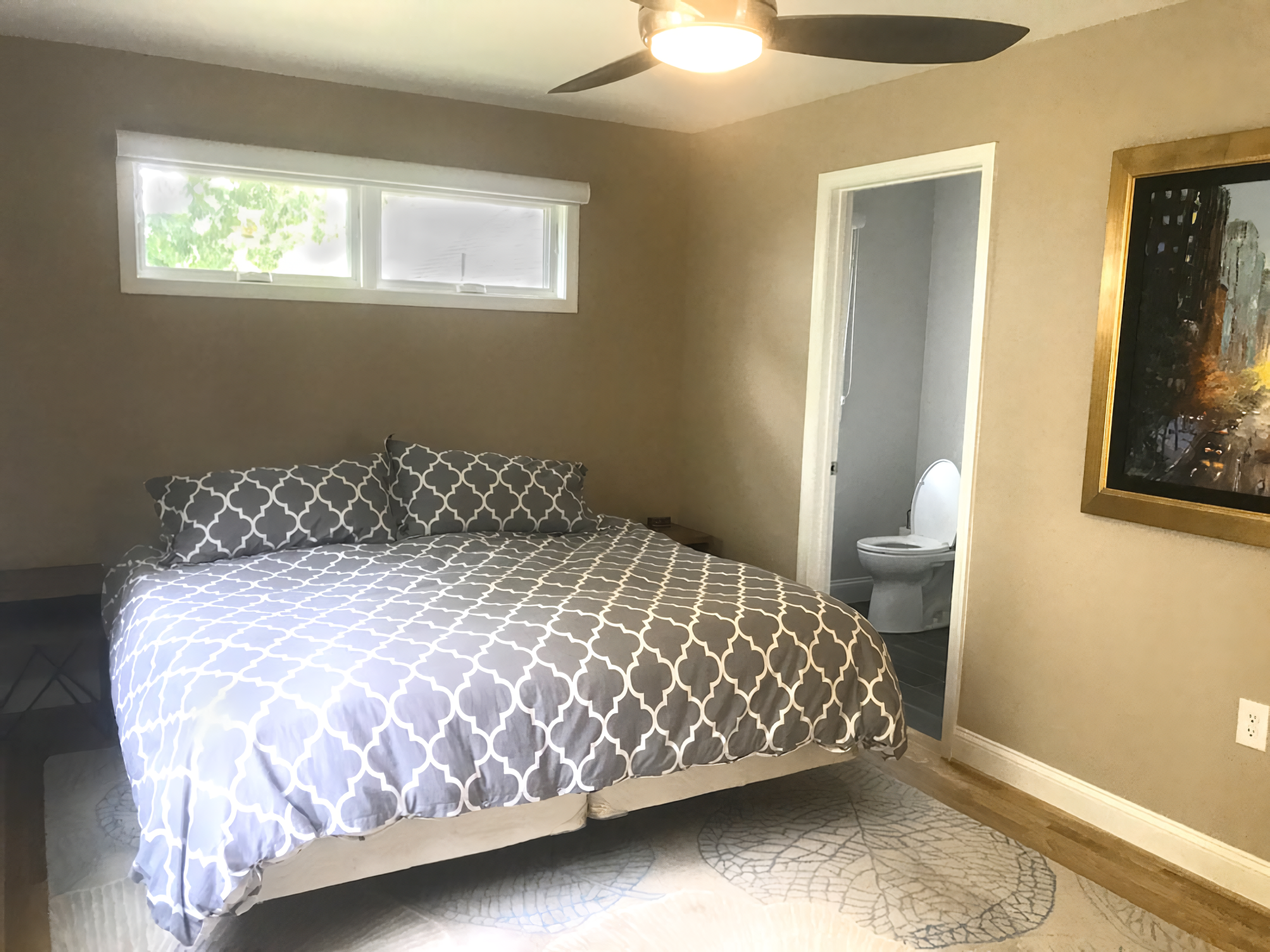 A bedroom with a bed covered in a gray and white patterned quilt, two matching pillows, a window above the bed, and a bathroom visible through an open door.