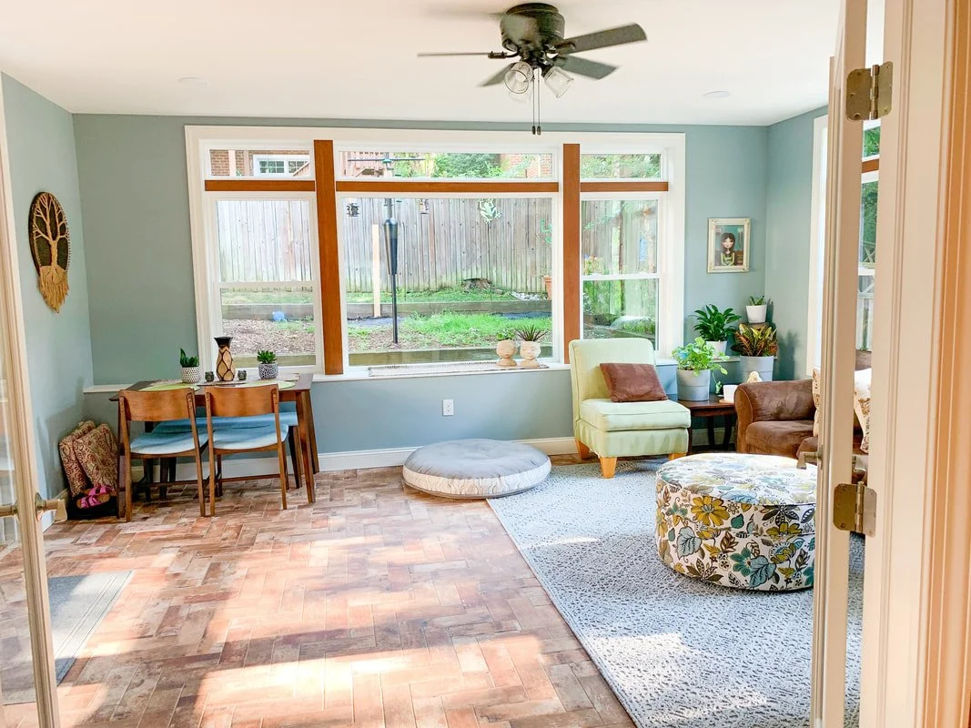 Bright living room with large front window, hardwood flooring, ceiling fan, green and brown chairs, an ottoman, and decorative indoor plants.