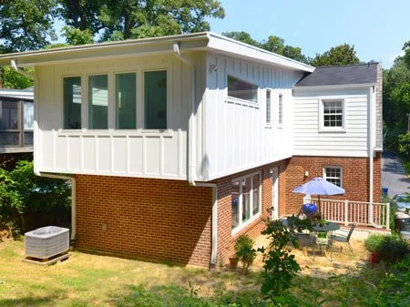 A two-story house with a brick base and white siding, featuring a screened porch on the upper level and an outdoor seating area with an umbrella on the lower level, surrounded by green trees.