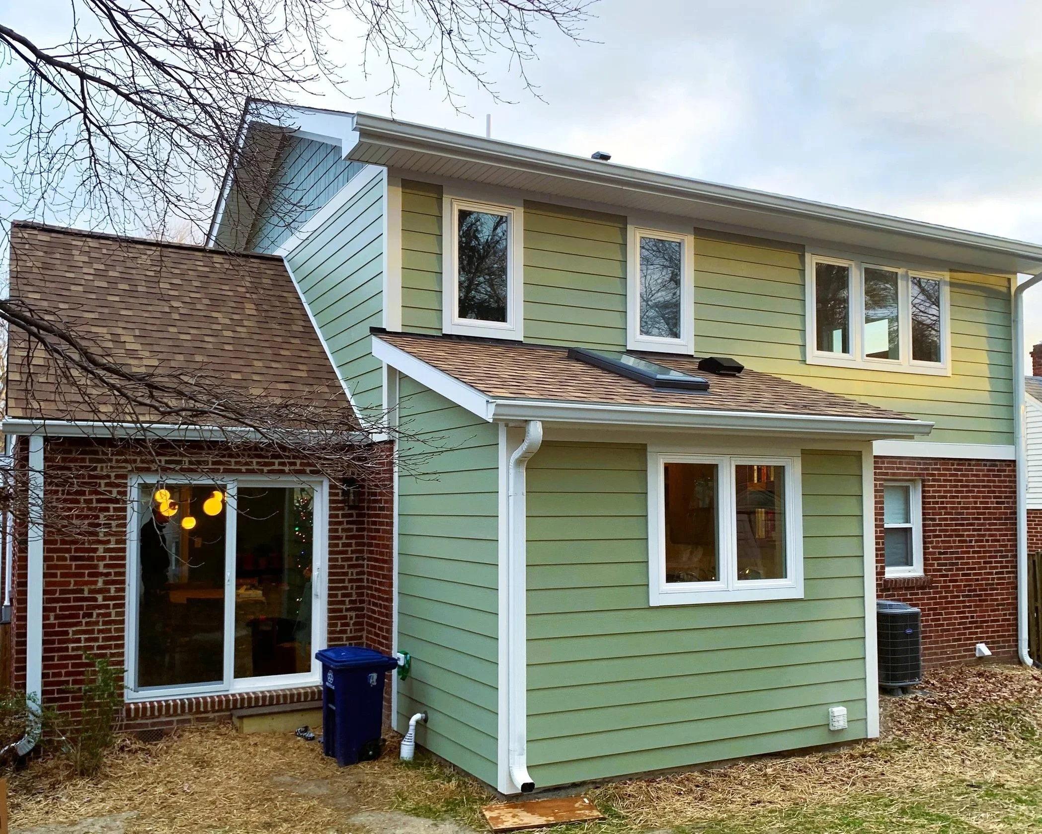 Front view of a two-story house with green siding and red brick exterior, multiple windows, and a small backyard with a blue trash bin and an air conditioning unit.