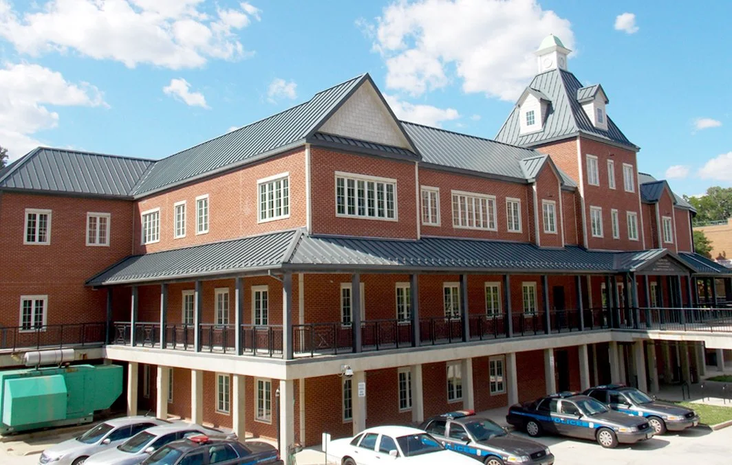 A large brick building with a metal roof, multiple windows, and a clock tower, with cars and police cars parked in front.