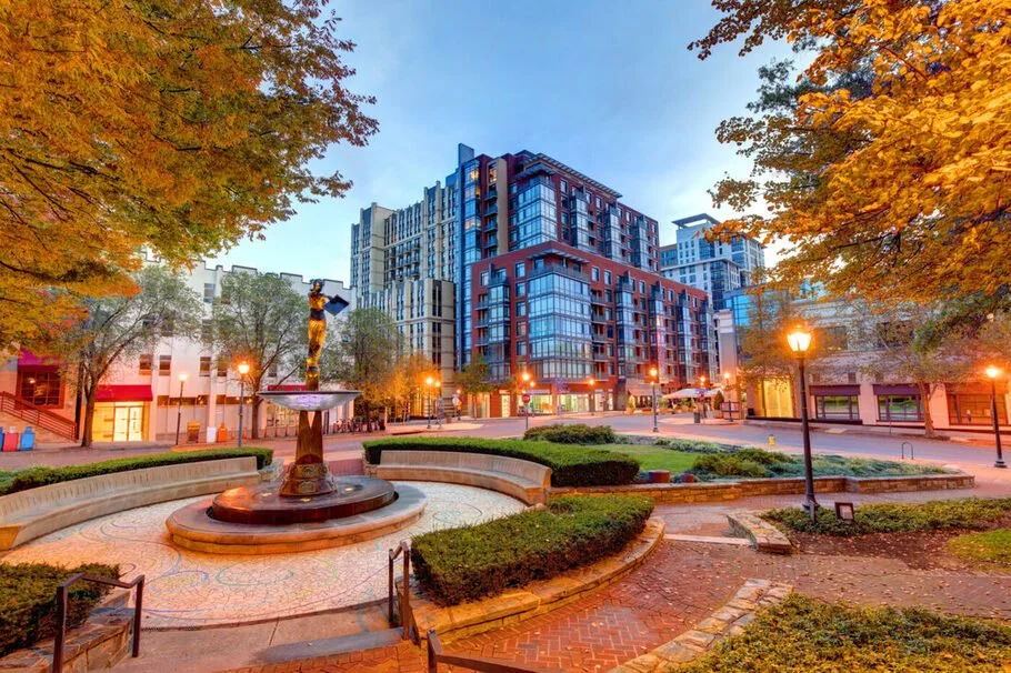 City park at dusk with a fountain and benches, surrounded by trees with orange leaves, and modern apartment buildings in the background.