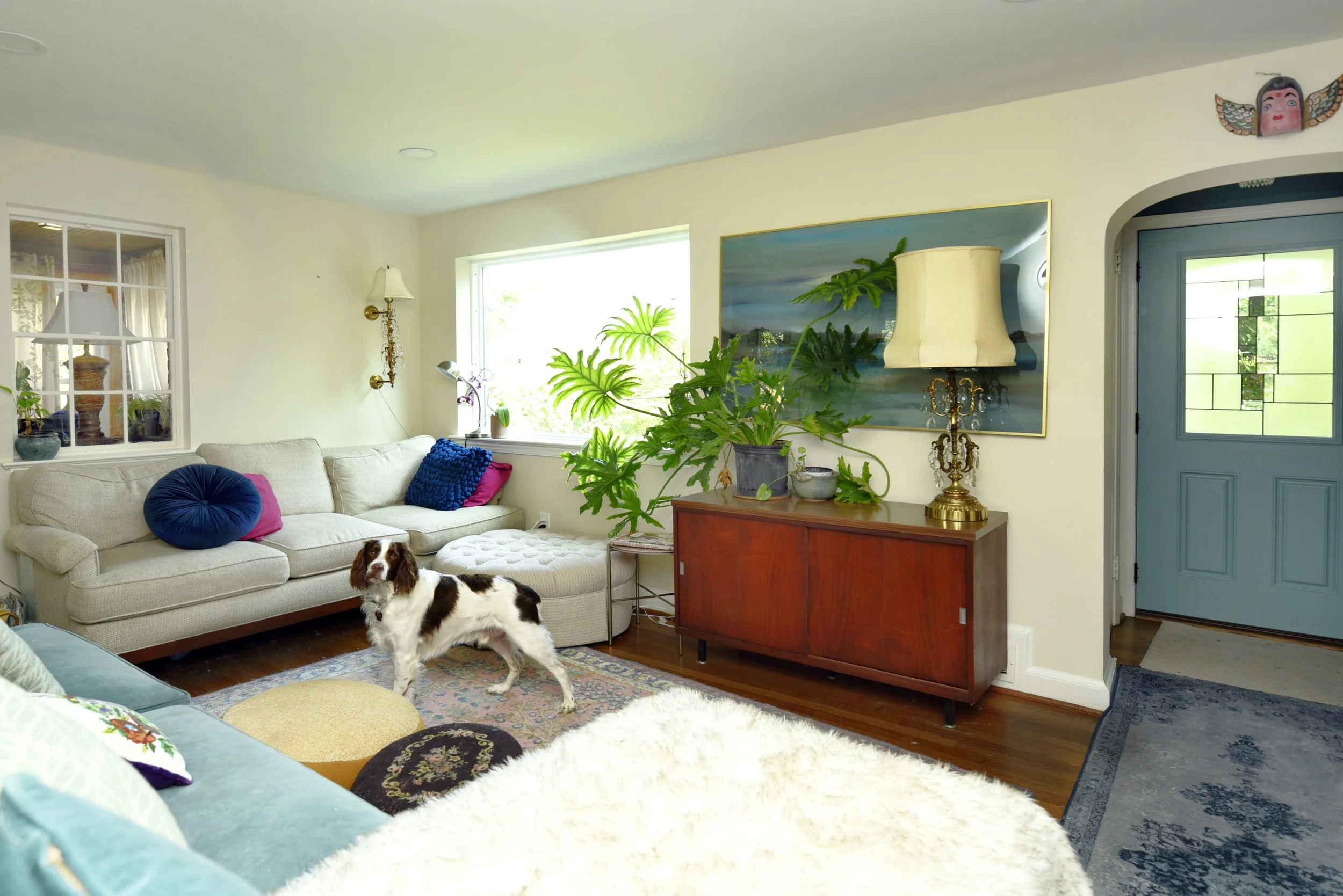 A living room with a cream sofa, colorful pillows, wooden floor, large window, and a dog standing near the sofa. There is a wooden sideboard with plants and a lamp, a painting on the wall, and an exit door next to an archway.