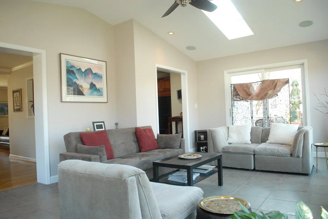 Living room with beige walls, skylight, ceiling fan, beige and gray sofas, red pillows, black coffee table, framed ocean landscape art, small black bookshelf, window covered with a sheer pink cloth, and mosaic tile flooring.