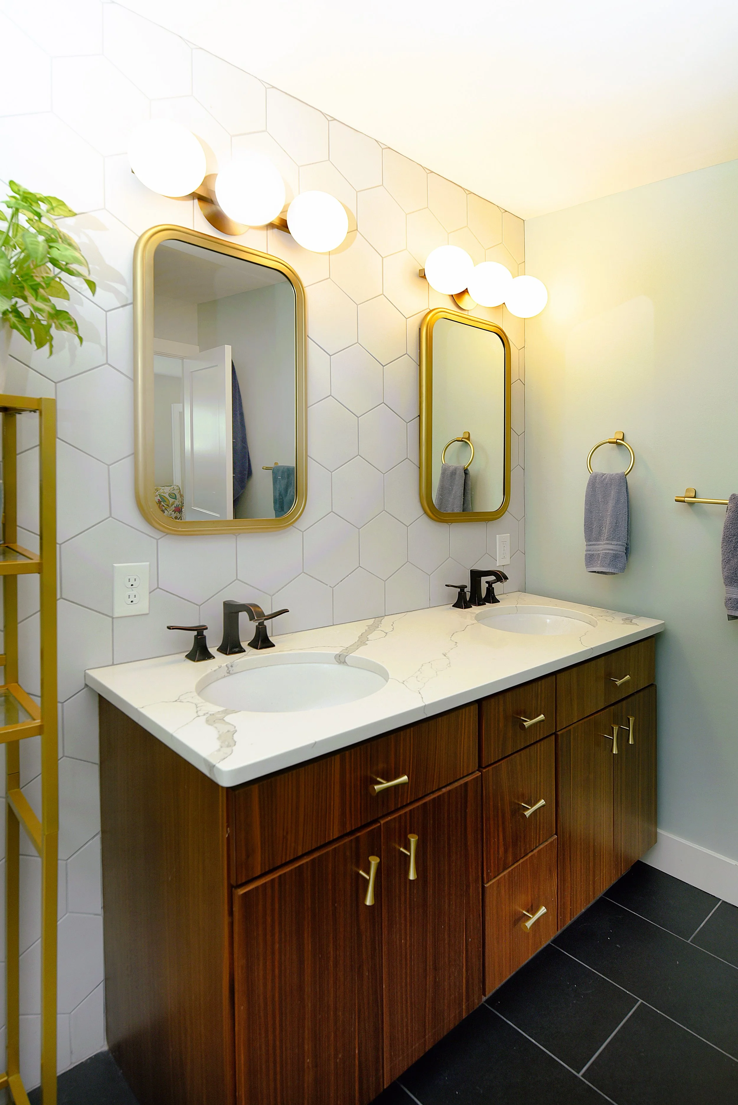 Bathroom with double vanity, wood cabinets, white marble countertop, black faucets, two oval mirrors with gold frames, hexagonal wall tiles, wall-mounted lighting, and gray towels on gold rings.