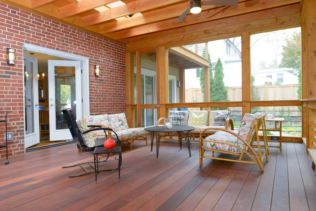 A covered porch with wooden ceiling and flooring, red brick wall, glass double doors, outdoor furniture including a loveseat, chairs, tables, and cushions, surrounded by a wooden railing and view of a backyard with trees and a wooden fence.