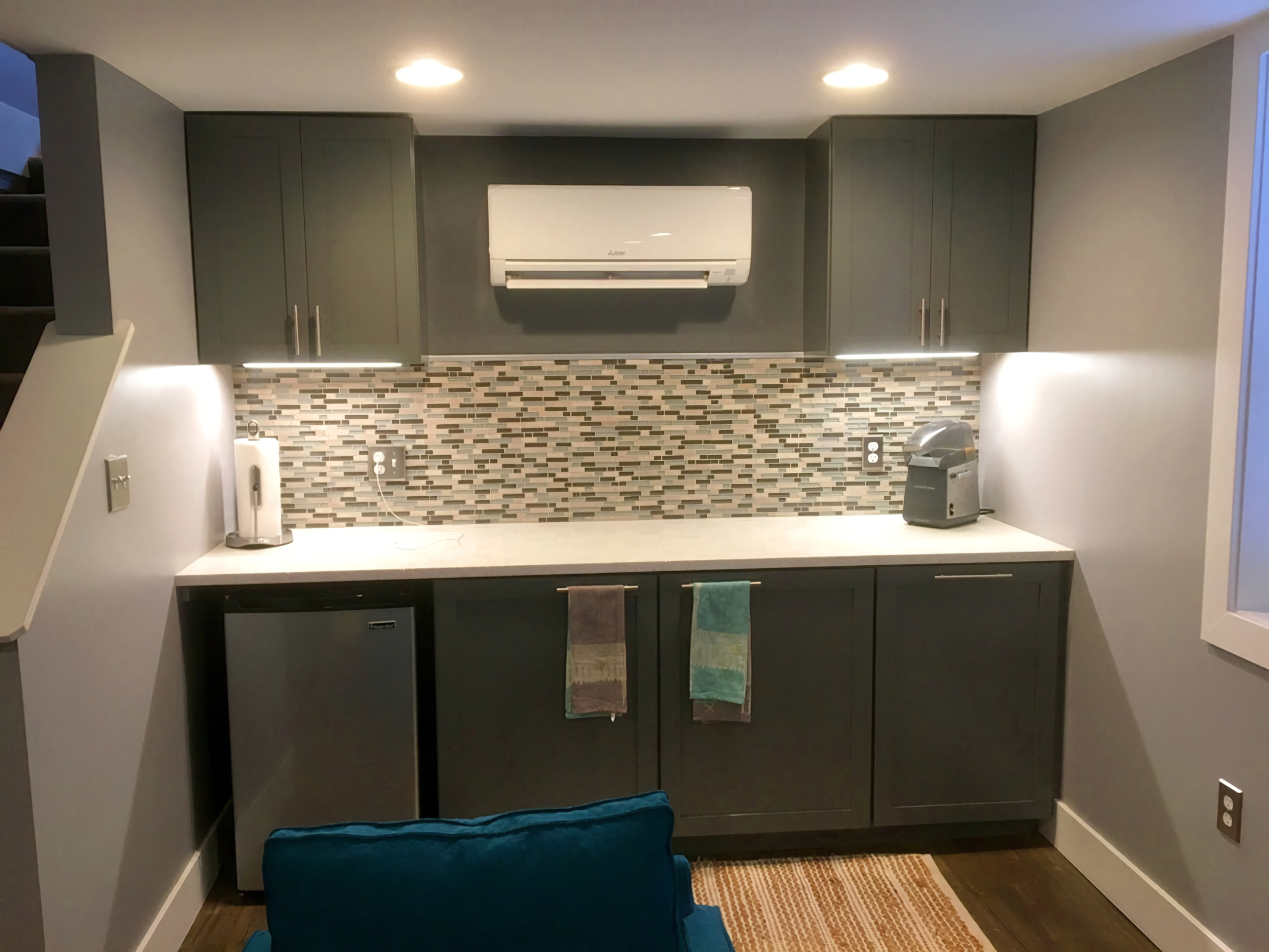 Small kitchen area with upper and lower dark gray cabinets, a tile backsplash, and appliances including a mini refrigerator, a coffee machine, and a paper towel holder. Overhead lighting illuminates the space.
