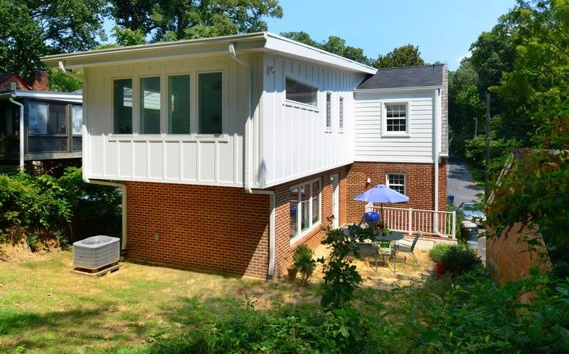 A two-story house featuring a brick lower level and a white upper level with large windows, surrounded by green trees and a backyard with outdoor furniture and an umbrella.