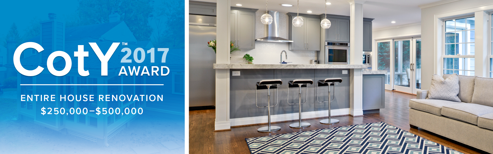 Interior view of a modern open-concept kitchen and living room with gray cabinets, a stainless steel oven and microwave, a marble island with three bar stools, a beige sofa, and a patterned rug near large windows and sliding doors.