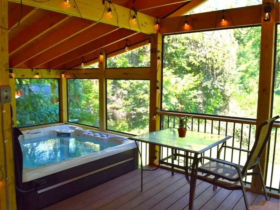 Sunlit porch with a hot tub, a glass table with a small potted plant, a chair, and lush green trees outside.