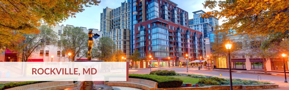 Cityscape view of downtown Rockville, Maryland, with modern high-rise buildings, trees with autumn leaves, street lamps, and a small park area.