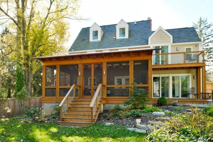 Back view of a multi-story house with a screened porch and a wooden deck, surrounded by trees and a garden.