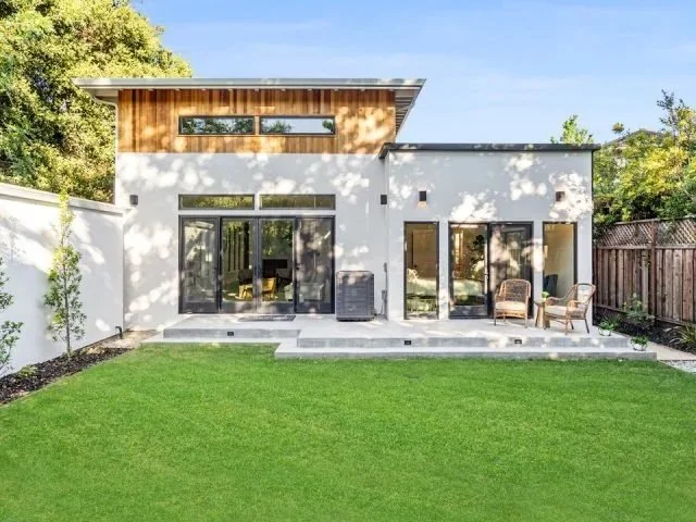 Modern backyard with a grassy lawn, concrete patio with outdoor furniture, and a white house with large glass doors and windows, wooden accents, and a fenced yard.