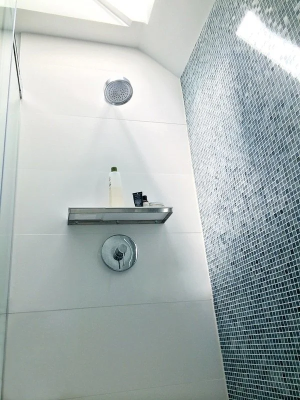 Shower with white tiles and a small metal shelf holding toiletries, viewed from a low angle.