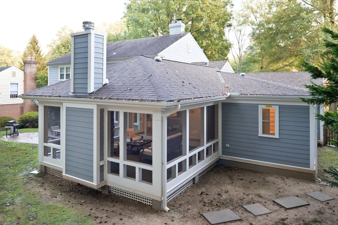 Backyard view of a two-story house with a screened porch, gray sunroom, and outdoor seating area, surrounded by trees.
