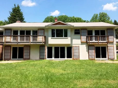 Two-story house with a green lawn, large windows, and balconies on the upper level, set against a blue sky with a few clouds.