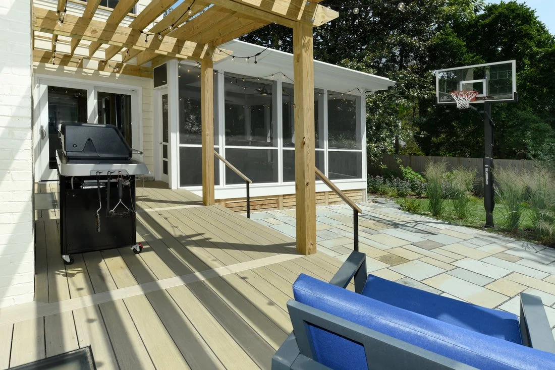 A backyard patio with a wooden deck under a pergola, a grill, a screened porch, seating, and a basketball hoop, surrounded by trees and plants.