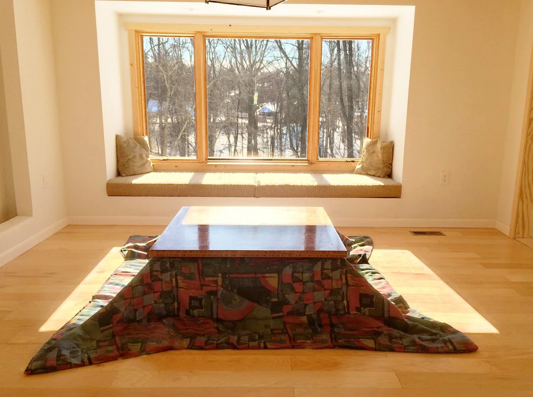 A cozy living room with a large window seat overlooking a winter landscape with trees and snow. There is a wooden coffee table in front of a patterned sofa on a light-colored wood floor.