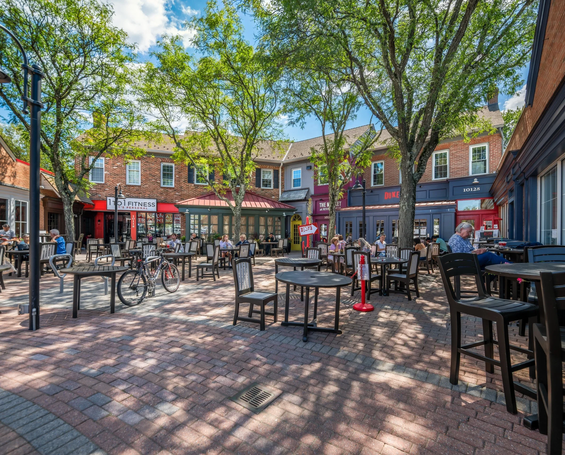 An outdoor shopping or dining area with tables and chairs, surrounded by buildings with brick and painted facades, trees providing shade, and people enjoying the setting on a sunny day.
