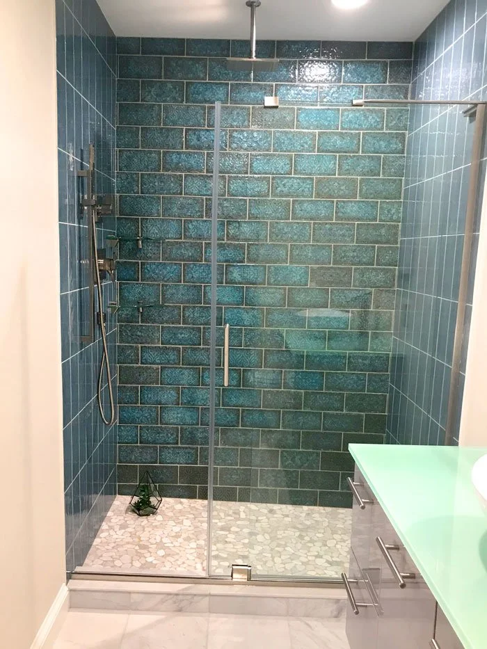 Modern walk-in shower with blue tiled walls, sliding glass door, white pebble shower floor, and a small green plant in a glass terrarium on the floor.