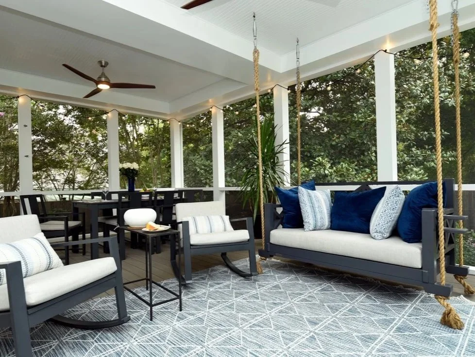 A screened porch with outdoor furniture, including two armchairs, a swing with multiple pillows, and a dining table with chairs, decorated with string lights and surrounded by greenery.