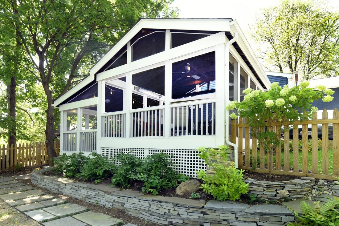 A screened-in porch with white framing and black screens, surrounded by lush greenery and a stone pathway.