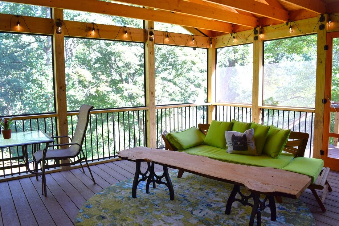 A screened-in porch with wooden beams and string lights, furnished with a green cushioned sofa with a cat face pillow, a wooden coffee table, and a glass table with a plant, overlooking lush green trees.