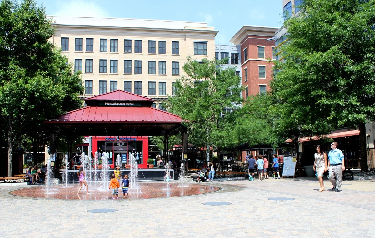 People playing in a fountain at Dawson's Market Stage in an urban plaza, with trees and multi-story buildings in the background.