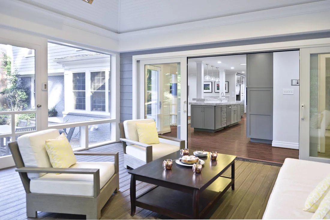 Sunlit indoor porch with white chairs, yellow pillows, and a dark wood coffee table in front of glass sliding doors leading to a kitchen with gray cabinetry and wooden floors.