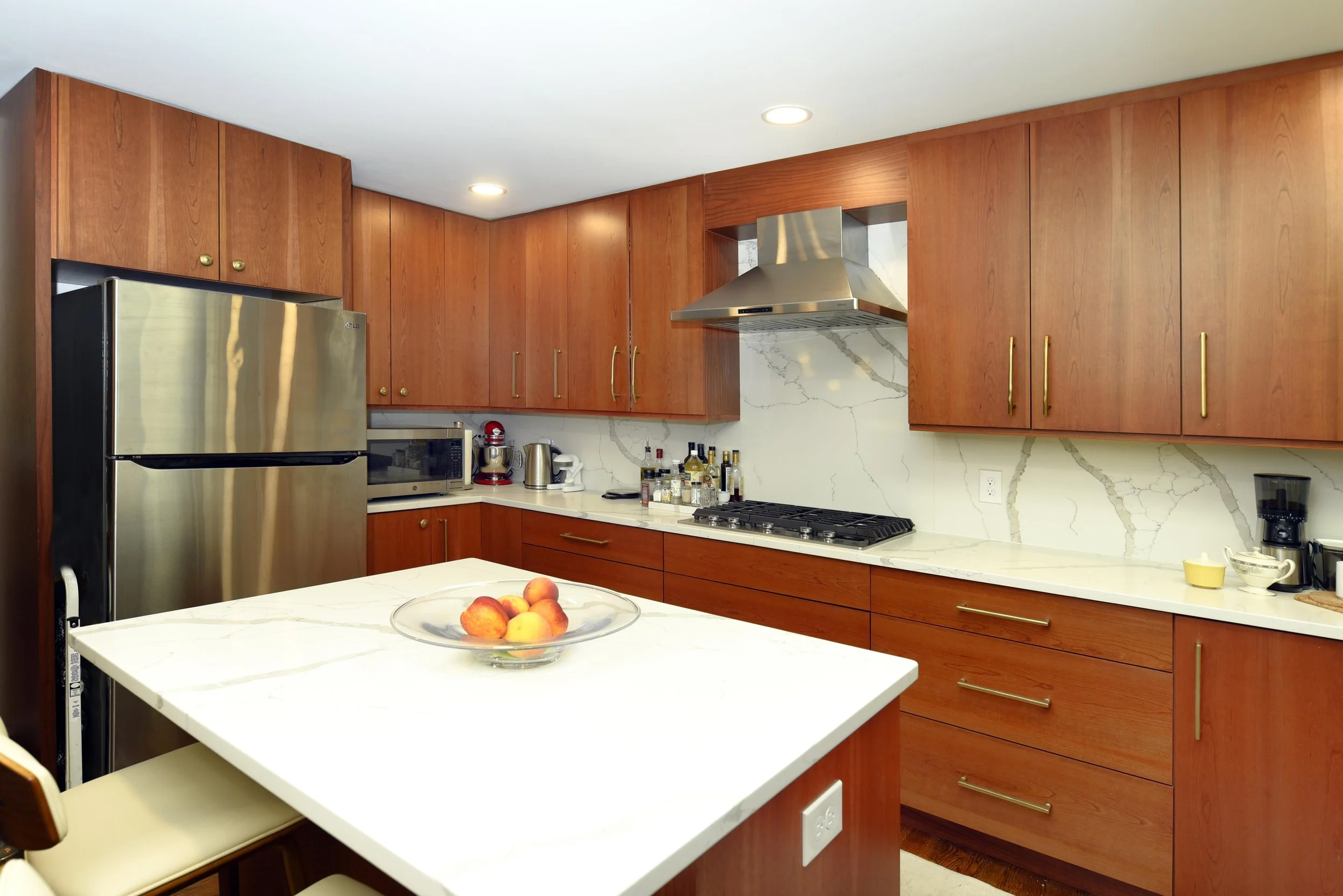Modern kitchen with wooden cabinets, white marble countertops, stainless steel appliances, and a kitchen island with a bowl of apples.