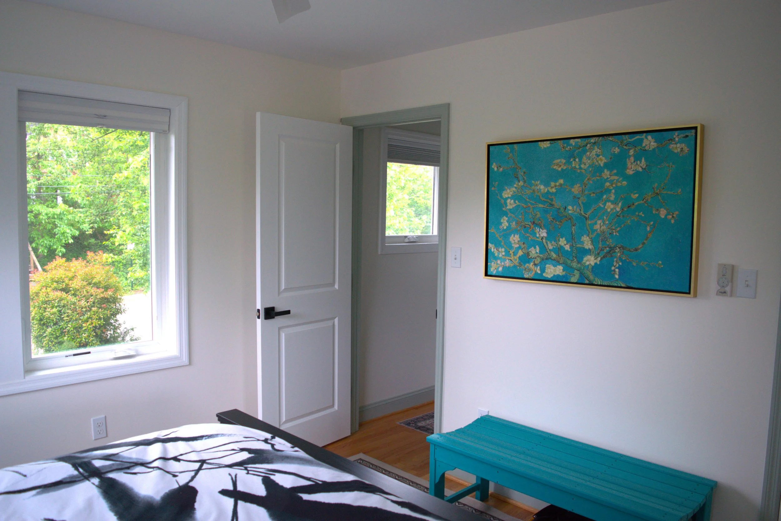 A bedroom with a window showing green trees outside, an open door with a white frame, a framed artwork of flowering branches on the wall, and a turquoise storage bench.