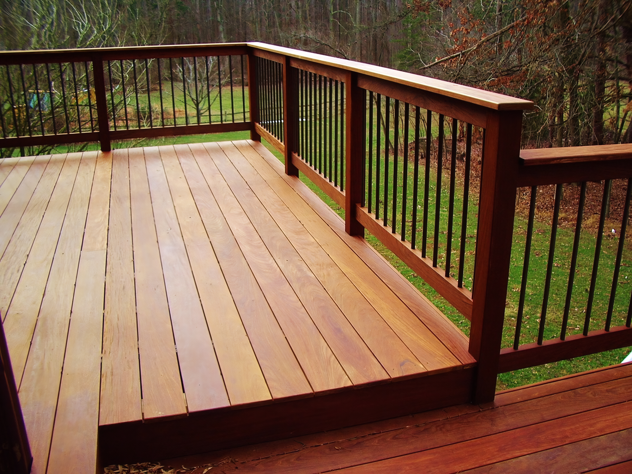 Wooden outdoor deck with railing overlooking a grassy yard and trees in the background.