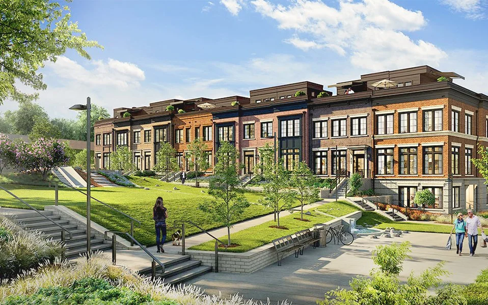 A row of modern townhouses with large windows and brick exteriors, surrounded by green lawns, trees, and a clear blue sky.