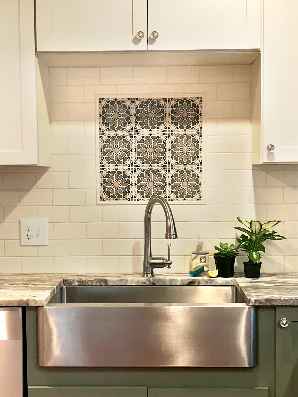 Kitchen sink with faucet, decorative tile backsplash, potted plants, cleaning spray, lemon wedge, electrical outlet, and beige cabinets.