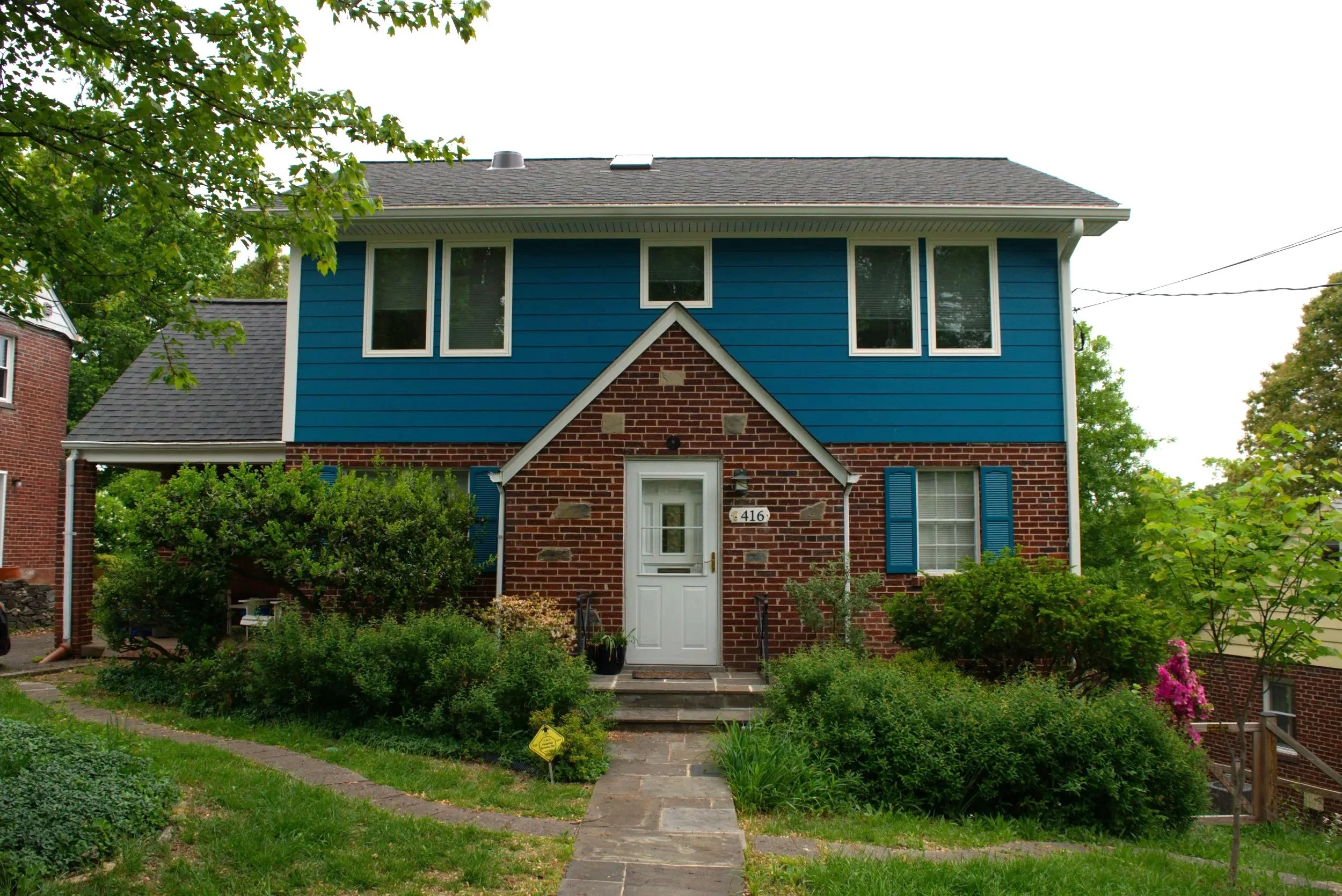 Front view of a house with a brick lower level and blue upper level, white door, and multiple windows, surrounded by greenery and trees.