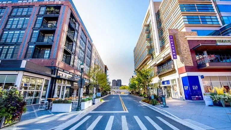 City street viewed from crosswalk, with modern buildings and storefronts on both sides, trees, and a blue sky