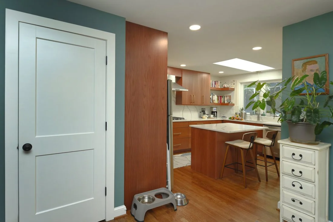 View of a modern kitchen with wooden cabinets, a small island with two chairs, a white side drawer with a large potted plant, and a colorful painting on the wall.