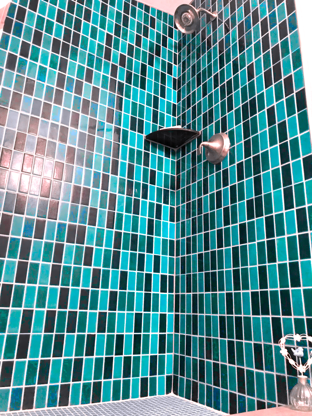 A corner of a shower with mosaic tiles in blue, turquoise, black, and white, a silver showerhead, a soap dish, and a small glass vase with a decorative string of beads and a small white shell.