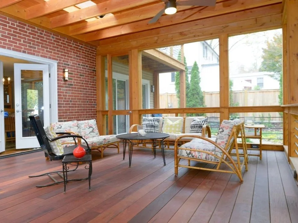 Covered porch with wooden ceiling, brick wall, glass door, and outdoor seating including a floral-patterned loveseat, rattan chairs, a black wire chair with a red vase, and a black round table, surrounded by a wooden railing and fenced yard.