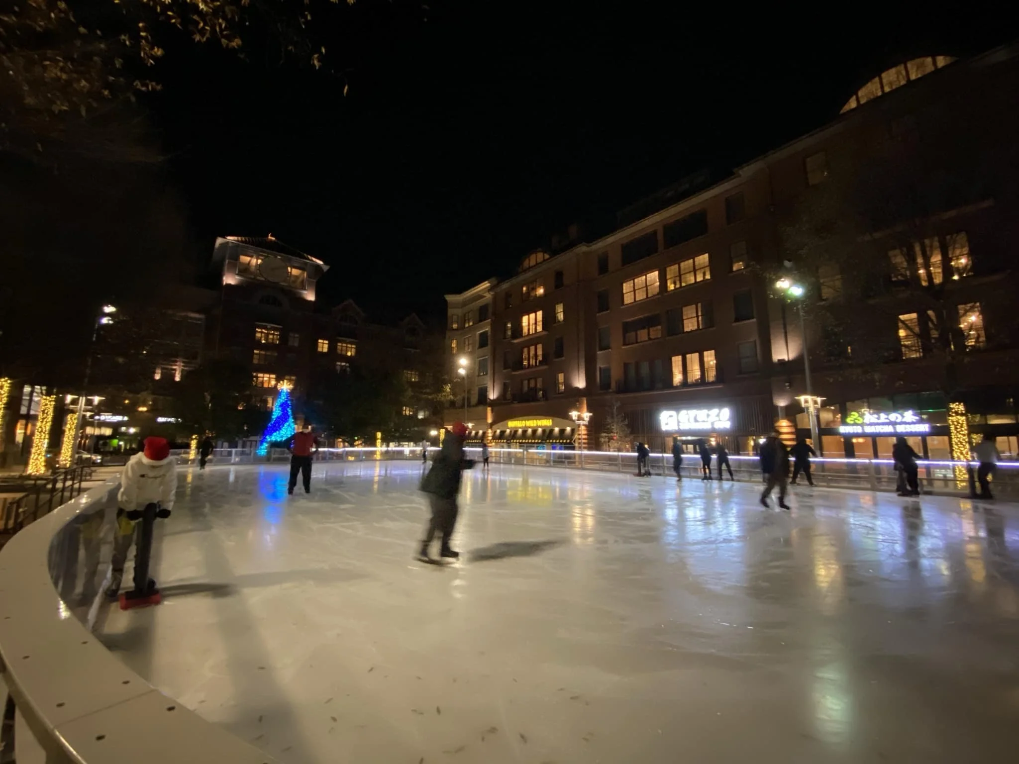 Night view of an outdoor ice skating rink with people skating, surrounded by lit trees and a building with lit windows and signs.